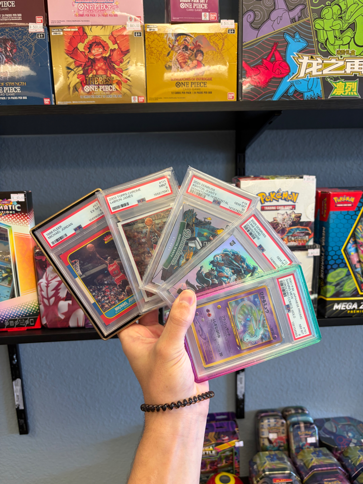 Stack of PSA graded Pokémon, Jordan, LeBron, and Jeanty slabs held up in front of the One Piece and Pokémon sealed wall